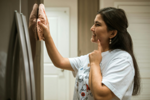 Imagem de uma mulher sorrindo enquanto limpa a casa com um pano e borrifador, representando leveza e praticidade na rotina doméstica.